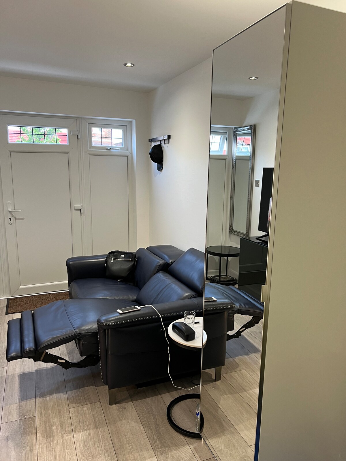 A comfortable black leather recliner is positioned next to a sleek mirror, reflecting the light from the white walls. A door with window panels leads outside, while a small table holds a phone charger. The flooring features light wood planks, enhancing the modern appearance.