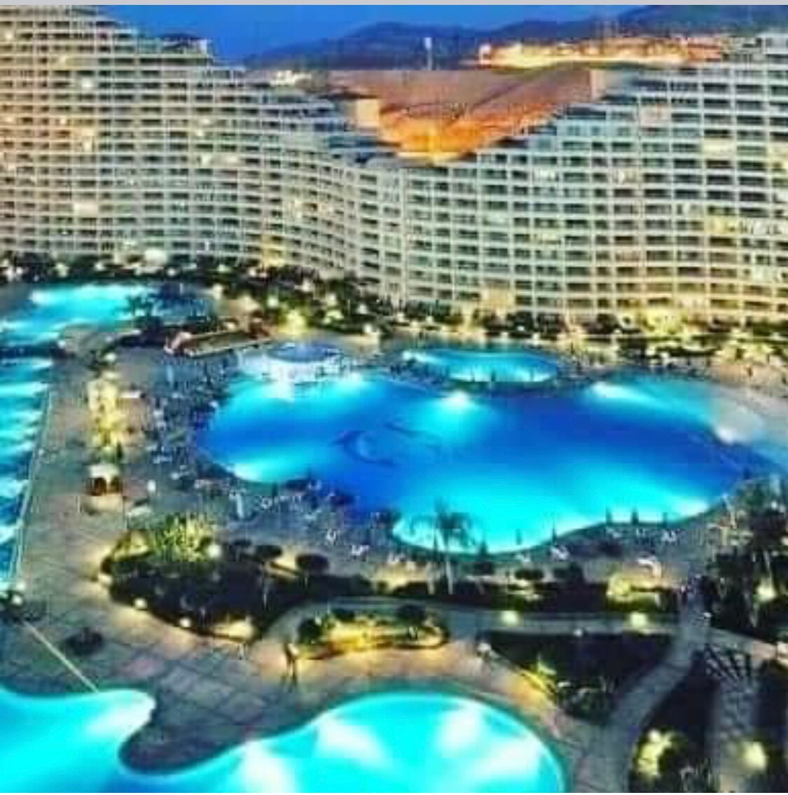 An expansive resort layout is visible, featuring multiple interconnected swimming pools illuminated in shades of blue at dusk. Surrounding landscaped areas include lounge chairs and palm trees, contributing to a relaxed environment. The buildings rise in the background, showcasing a modern architectural design.