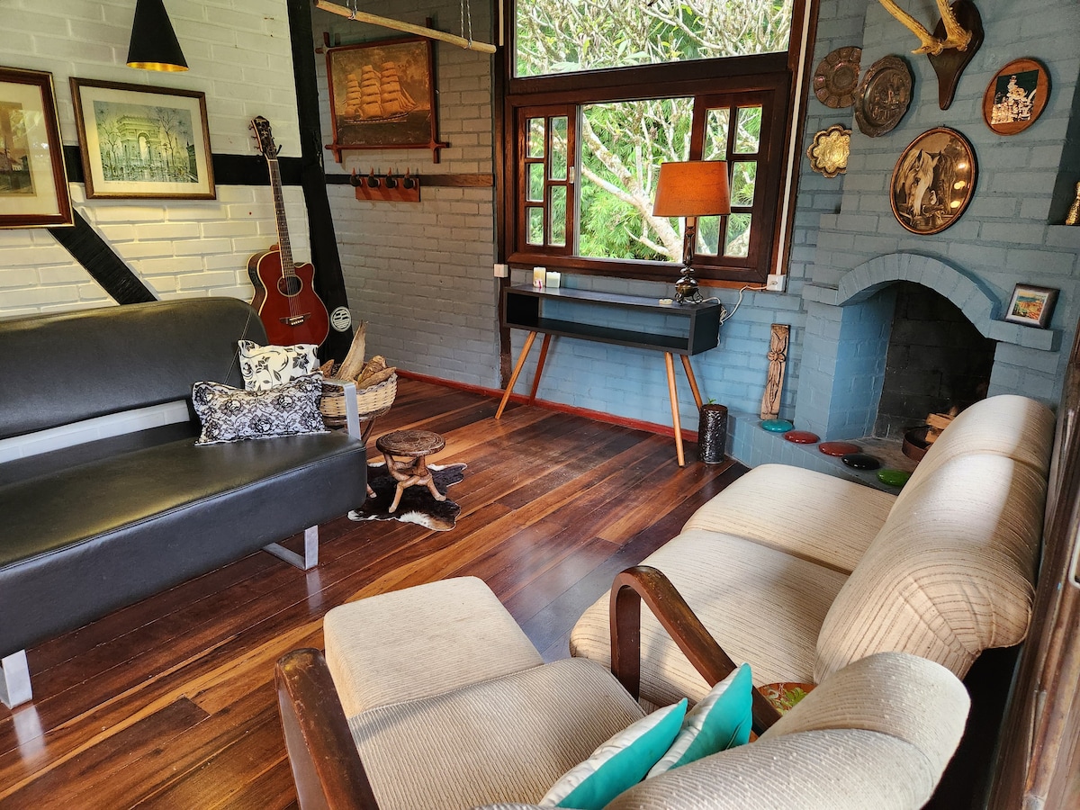 A comfortable seating area features a dark leather sofa and a light-colored upholstered sofa, complemented by wooden accents. Decorative items, including framed artwork and a guitar, adorn the walls. A small side table and a lamp provide additional warmth to the cozy atmosphere.