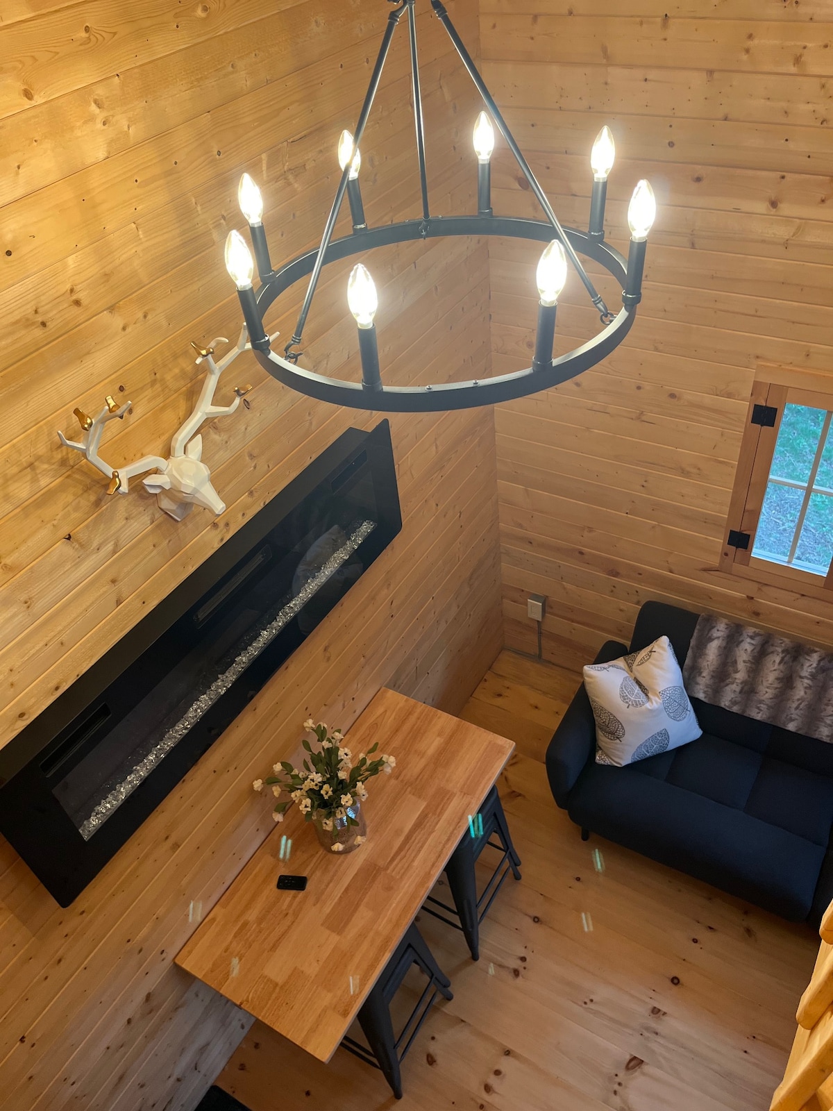 An overhead view of a cozy interior features warm wood paneling and a modern chandelier. A small table with two stools sits beneath a decorative wall-mounted antler, while a comfortable dark sofa is positioned nearby, accented by a patterned throw pillow.