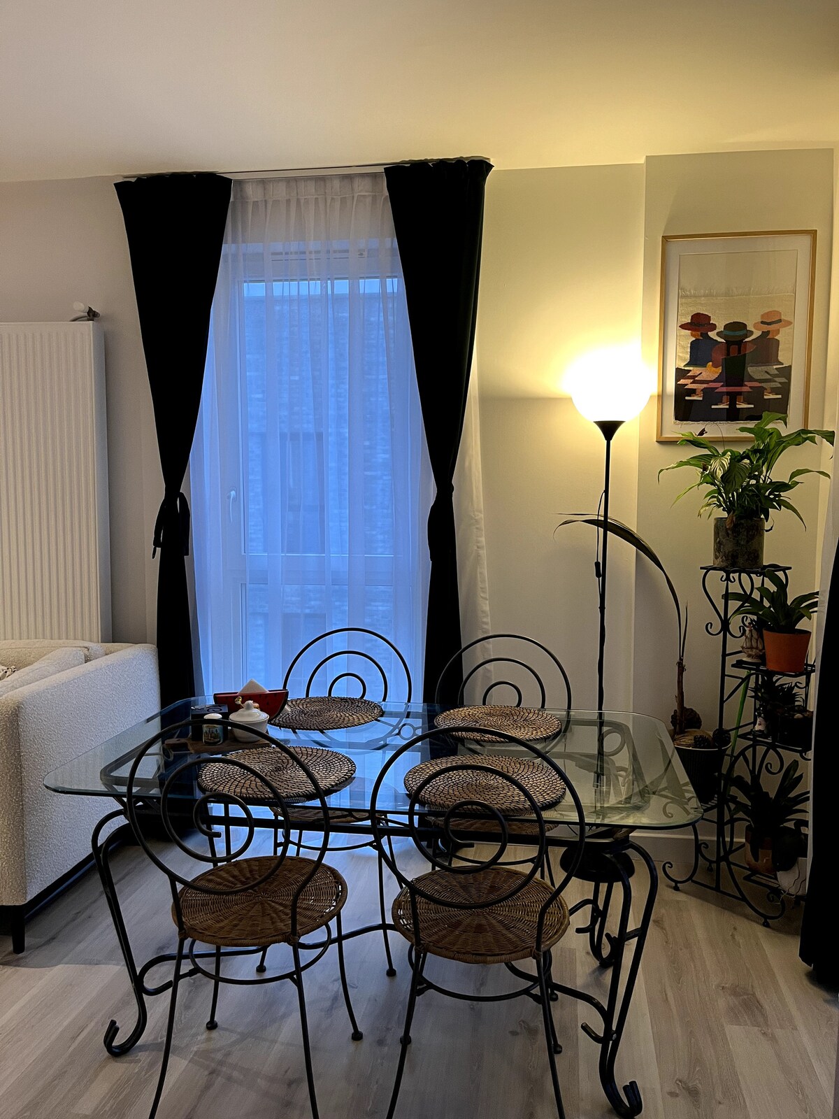 A small dining area includes a glass table surrounded by four metal-framed chairs with woven seats. A tall lamp stands beside the table, illuminating the space. A window is partially covered by light curtains, and a framed artwork is displayed on the wall nearby.