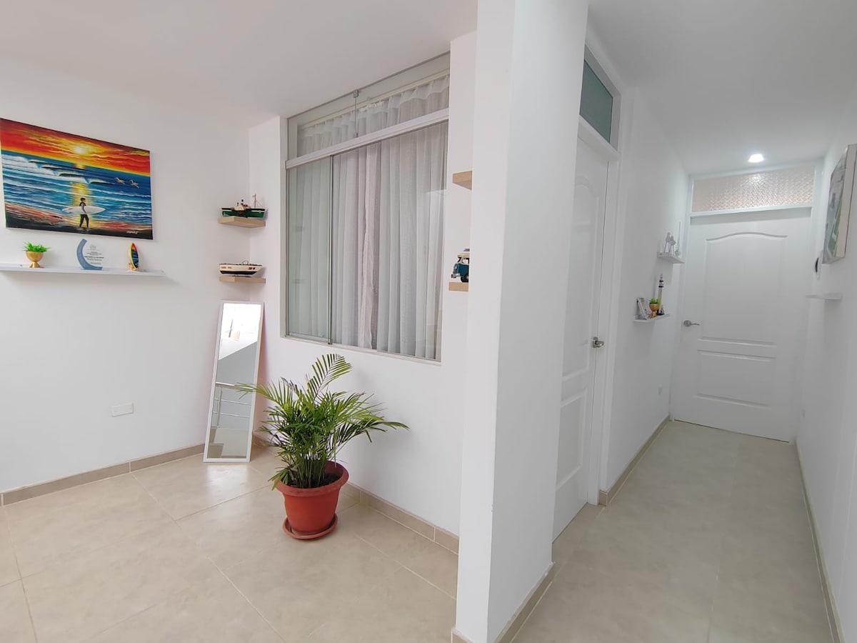 A bright corridor features white walls and a neutral tiled floor. Large windows allow natural light to fill the space, with a small potted plant placed beside a wall mirror. Colorful artwork hangs on the wall, adding a touch of vibrancy.