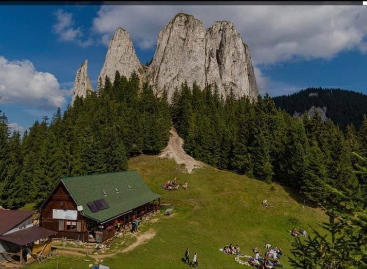 A rustic wooden cabin sits on a grassy area surrounded by dense coniferous trees. Tall, rocky peaks rise majestically in the background, and several groups of people are seen enjoying the outdoor space, some seated on the grass.