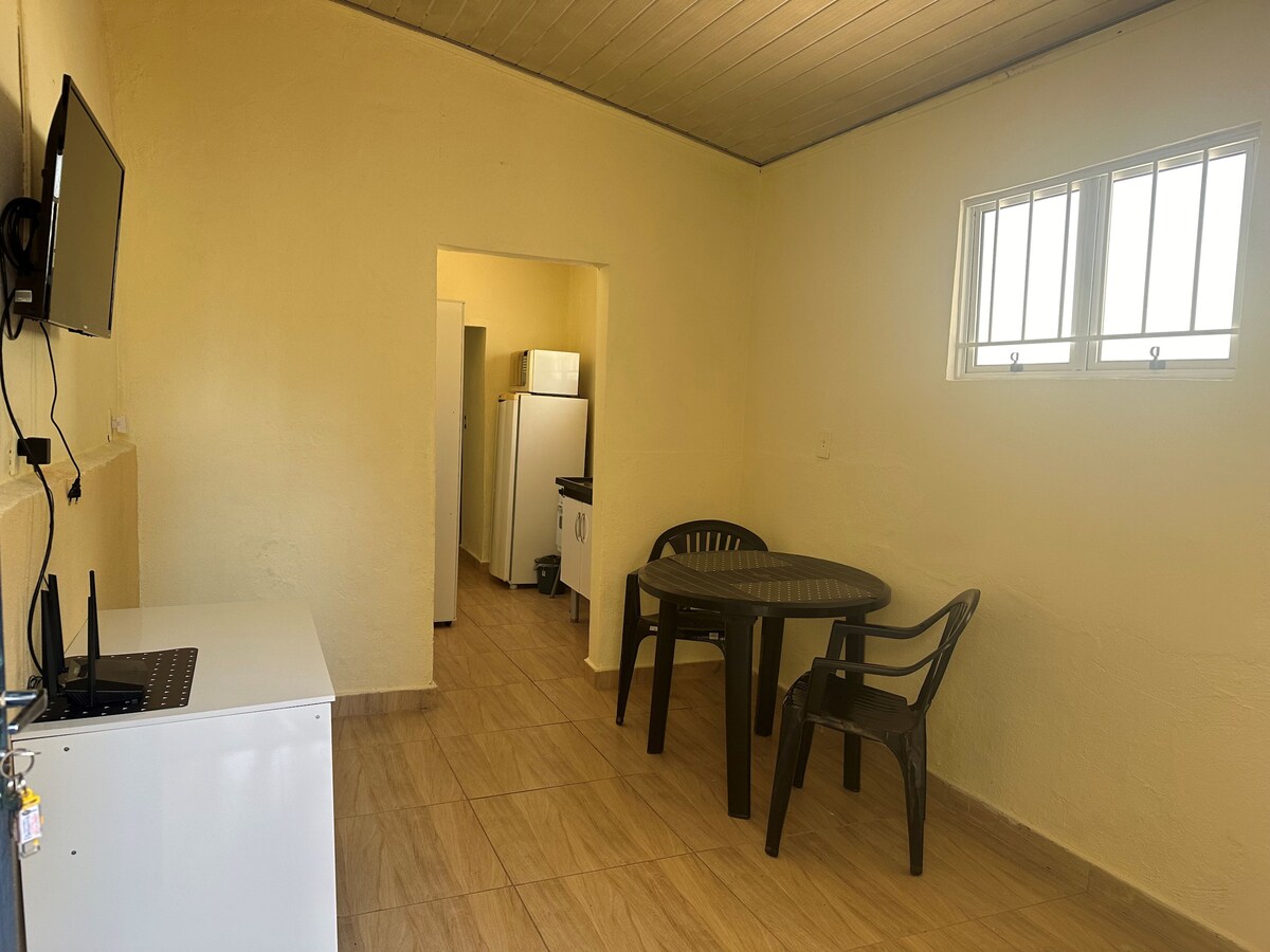 A simple dining area is shown with a round table surrounded by four black plastic chairs. A small refrigerator is visible beside the entrance to a kitchen area, which includes a stove and white cabinetry. Light filters through the two small windows.