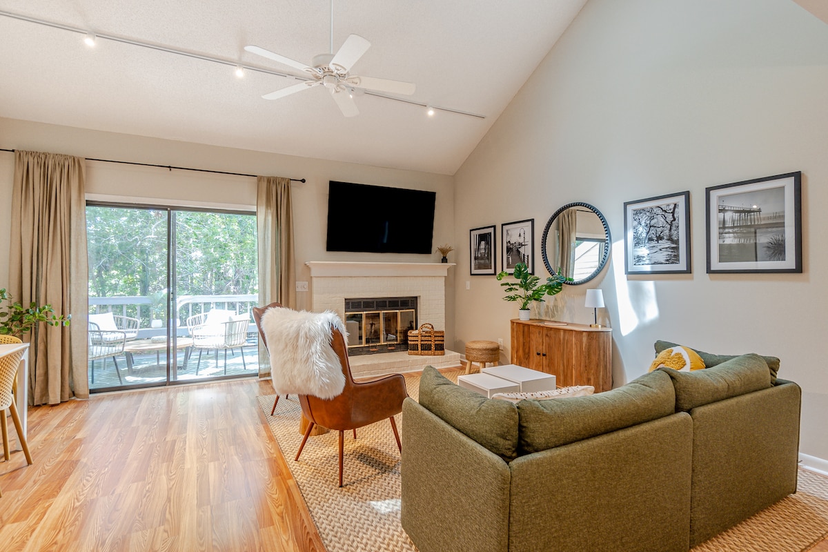 A bright and airy living space is showcased with large glass doors leading to a patio. Comfortable seating is arranged around a cozy area, featuring a sofa and accent chairs. Natural light floods the room, highlighting a simple decor with framed photographs on the wall.