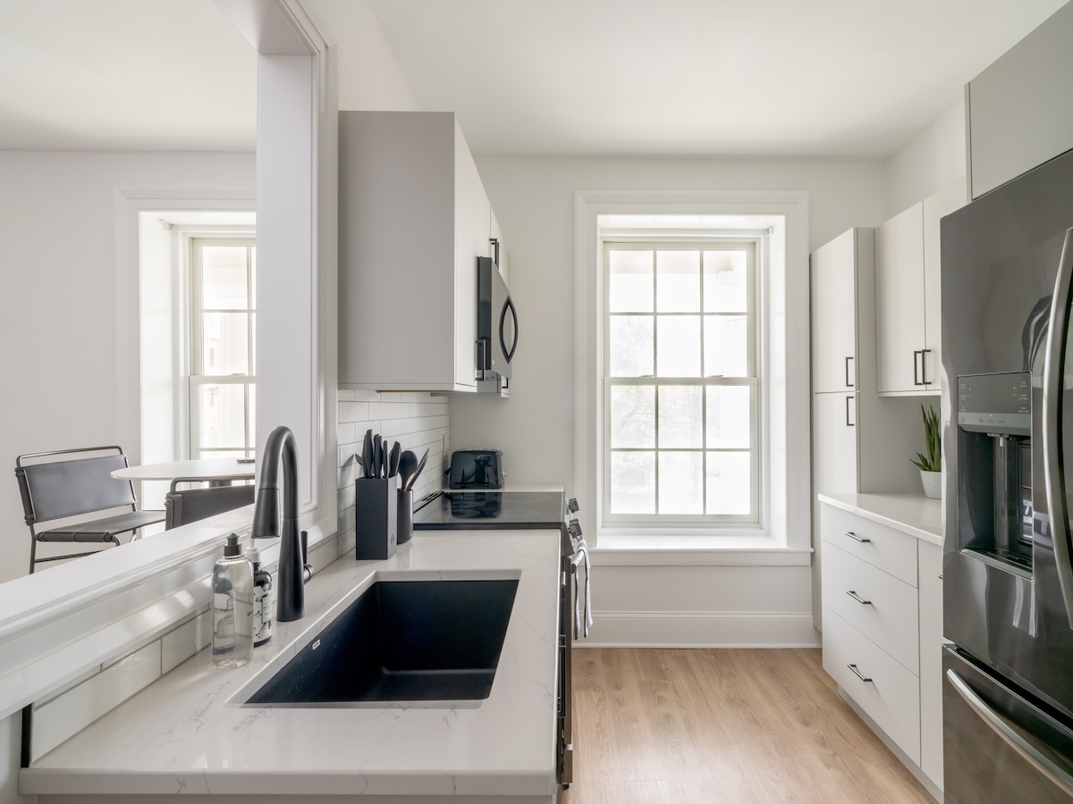 The kitchen features modern cabinetry with sleek hardware and a spacious black sink. A full-size refrigerator and stove are included among the black stainless-steel appliances. Natural light fills the space through a large window, highlighting the white countertops and light wood flooring.