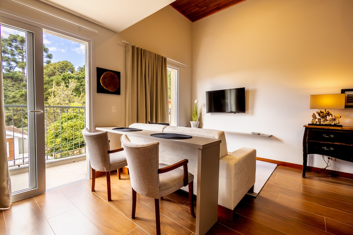 A well-lit living area is presented, featuring a comfortable sofa paired with two chairs around a small table. Natural light pours in through the glass doors, which lead to a private balcony. A flat-screen TV is mounted on the wall, enhancing the modern ambiance.