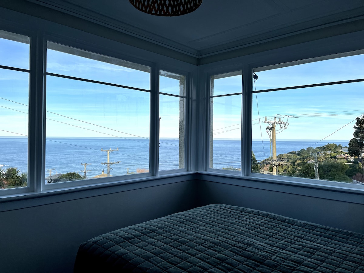 A bright bedroom features large windows that offer a panoramic view of the ocean and surrounding areas. The space contains a spacious bed topped with a quilted cover. Natural light fills the room, enhancing the sense of openness and tranquility.