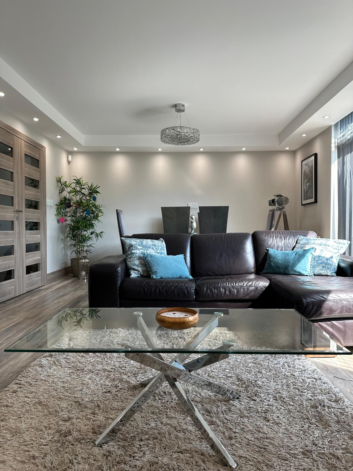 The living area features a comfortable leather sofa accented with decorative cushions, a glass coffee table placed on a plush area rug, and a potted plant for a touch of greenery. Soft lighting enhances the modern decor, complemented by a dining table visible in the background.