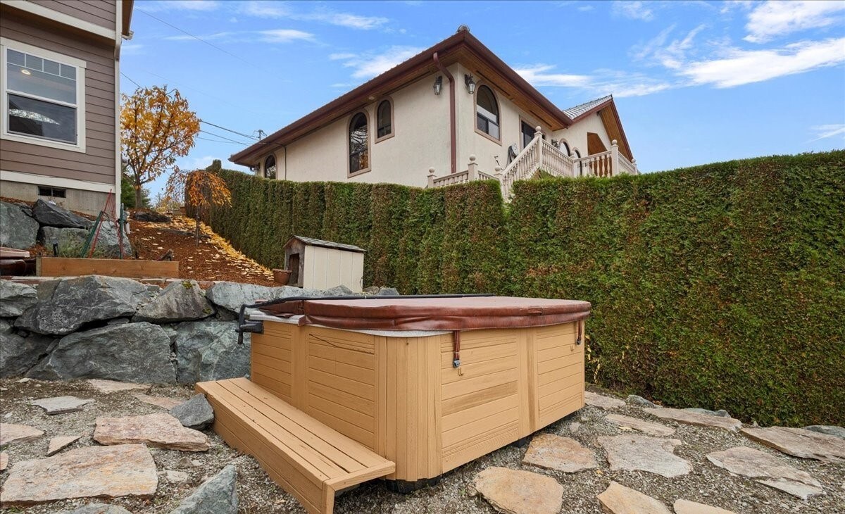 A well-maintained hot tub is positioned on a stone patio, surrounded by a neatly trimmed hedge. The tub’s cover rests on top, while a small wooden step leads up to it. A view of the home's exterior can be seen in the background.