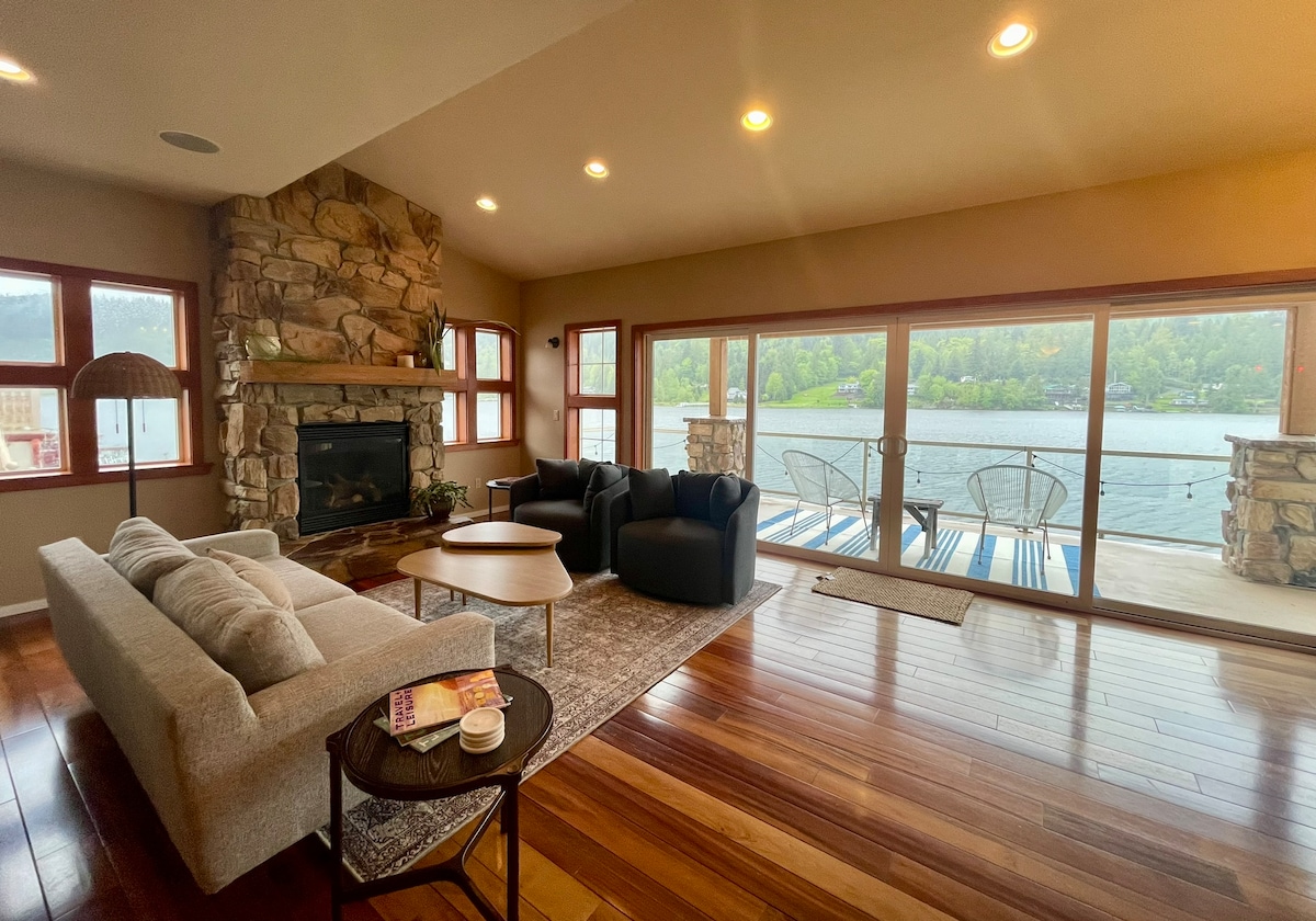 A spacious living area features large windows offering views of the lake. Comfortable seating arrangements include a light-colored sofa, two dark chairs, and a side table with a drink and reading material. A stone fireplace adds a cozy element to the room.