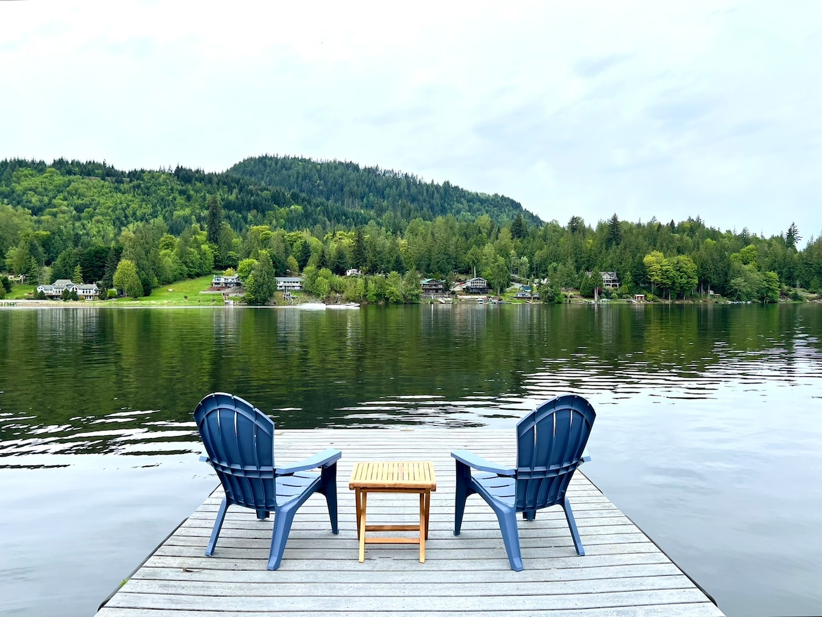Two blue Adirondack chairs are positioned on a lakeside dock, facing calm water and lush greenery on the opposite shore. A small wooden table sits between the chairs, creating a serene spot for relaxation and reflection.
