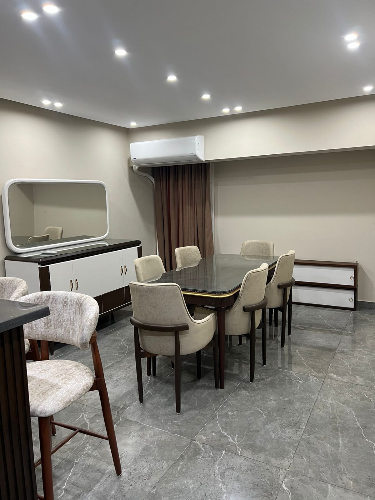 A dining area features a long, dark table surrounded by six cream-colored upholstered chairs. A mirrored cabinet adds elegance to the space, with a neutral color palette of walls and flooring. Soft ceiling lights enhance the inviting ambiance.