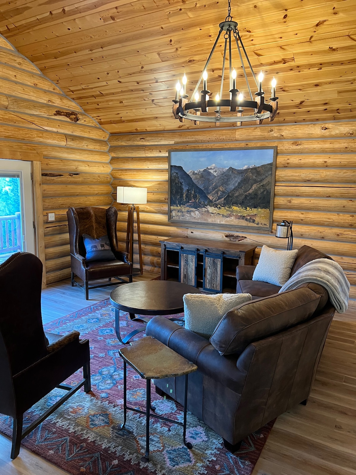 A cozy living area features log walls and a high ceiling. Comfortable seating includes a leather sofa and two wingback chairs. A round table sits at the center, surrounded by natural light from the nearby door. A landscape painting adorns the wall, adding visual interest.