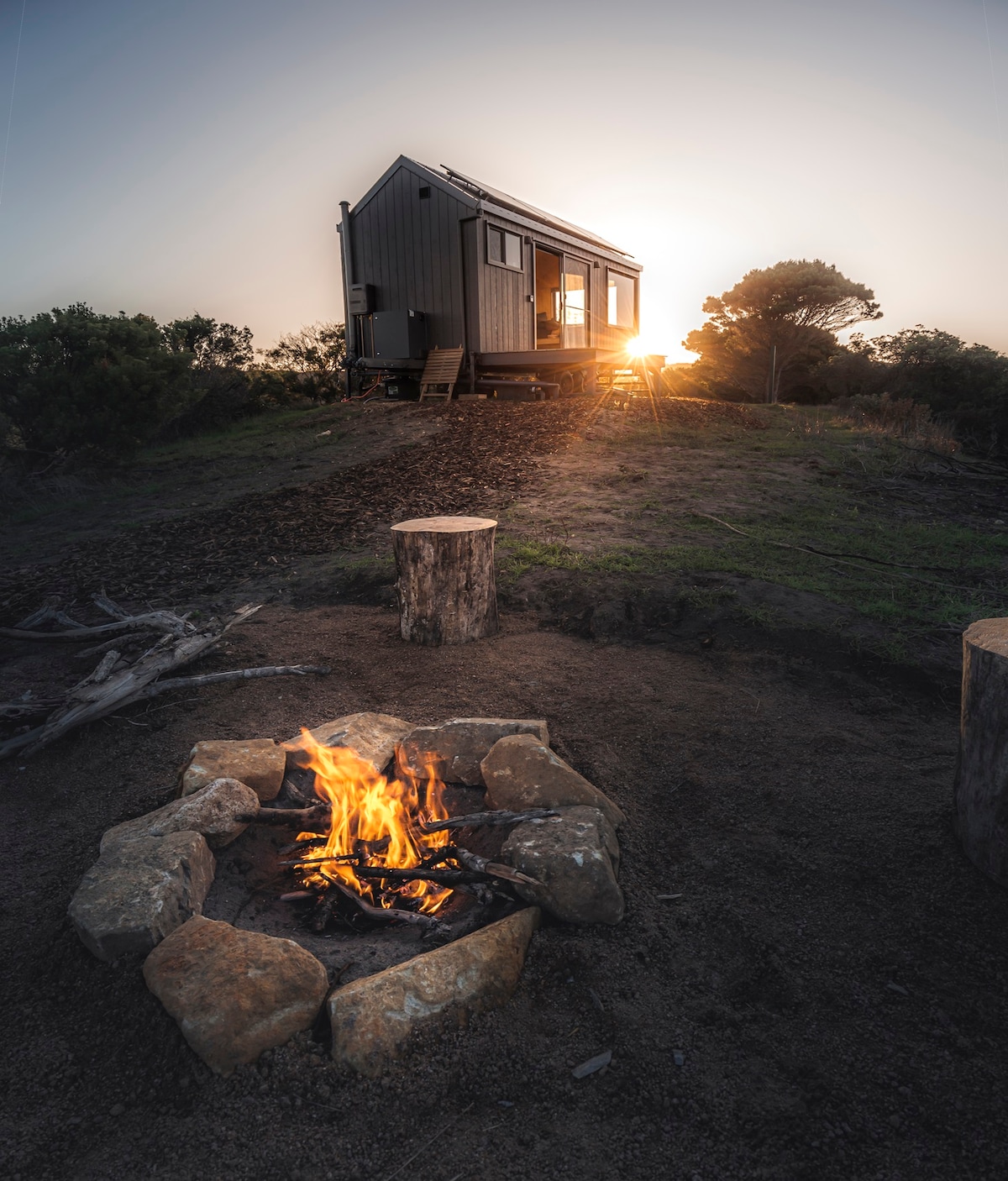 A cabin sits atop a grassy hill at sunset, casting warm light through its open doors. A stone fire pit is positioned nearby, surrounded by wooden stumps for seating, creating a cozy space to enjoy the evening.