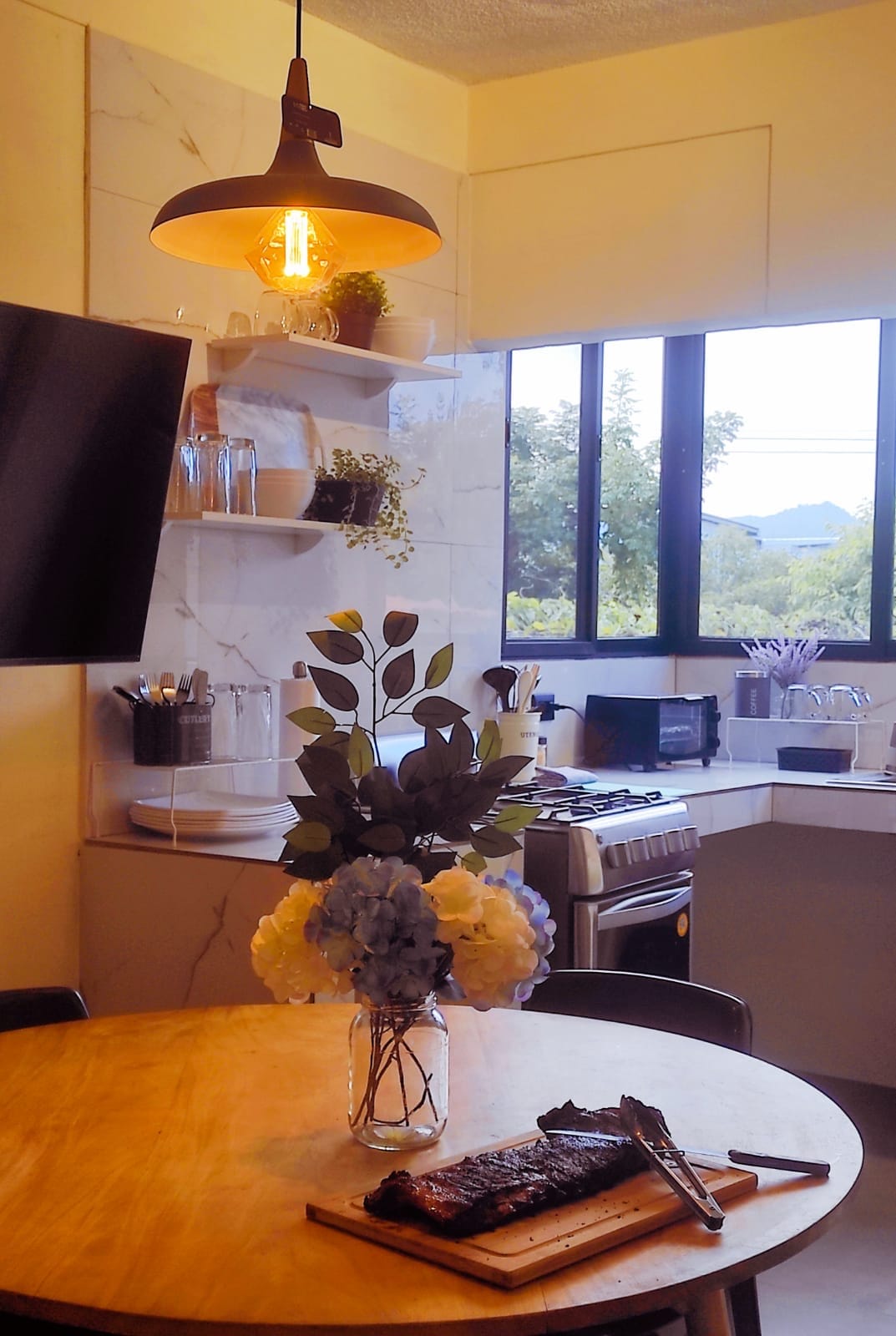 A modern kitchen is featured with a round wooden table at the center, adorned with a vase of flowers. A TV is mounted on the wall opposite, while shelves hold kitchenware. A window offers views of greenery, and a stove is visible in the cooking area.