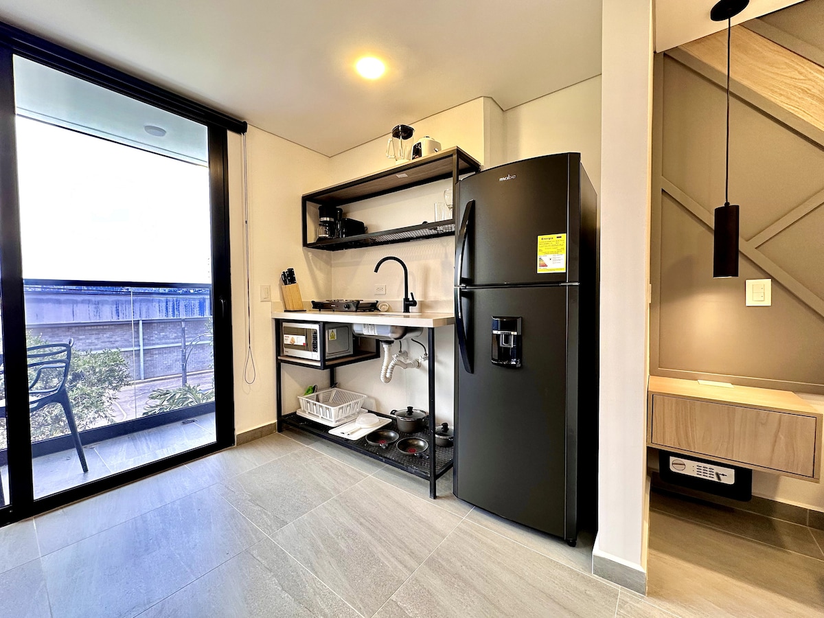 A modern kitchen is presented, featuring a sleek black refrigerator and stainless steel shelves. A sink and stove are visible, alongside essential kitchen appliances. Large glass doors lead to an outdoor area, allowing natural light to illuminate the space.