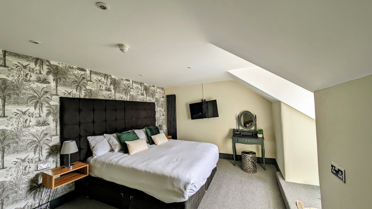A spacious room features a large upholstered bed adorned with decorative pillows. A wall-mounted television is positioned above a stylish dressing table. Light-colored walls are complemented by patterned wallpaper, while a sloped ceiling enhances the room's character.