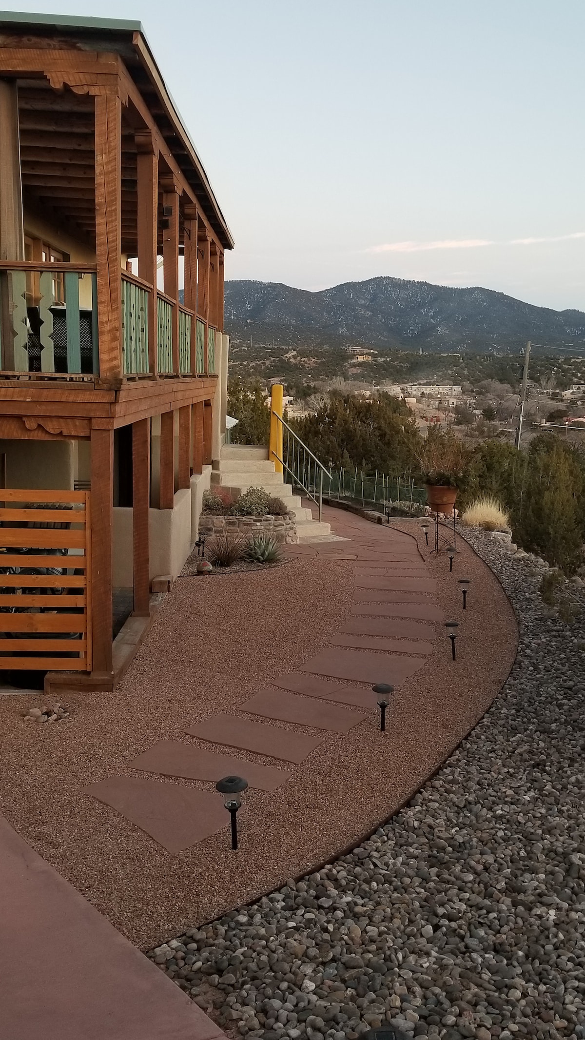 A landscaped pathway, lined with low garden lights, winds its way along a gravel base towards a two-story home with a wooden balcony. Soft mountains are visible in the background, creating a serene backdrop as the sun sets.