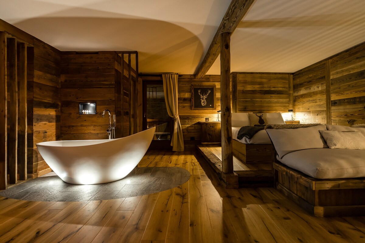 An elegantly designed open space features a freestanding bathtub at its center, surrounded by wooden walls and a warm wood floor. A cozy sleeping area is located in the corner, with soft bedding and subtle lighting enhancing the serene ambiance.