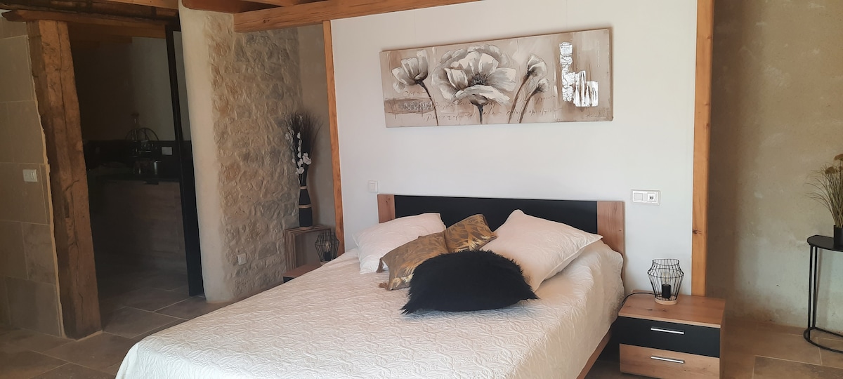 A spacious bedroom presents a comfortable bed adorned with soft pillows and a textured throw. A piece of floral wall art hangs above the bed, while wooden beams and natural stone accents enhance the room's character. Gentle lighting is provided by a bedside lamp.