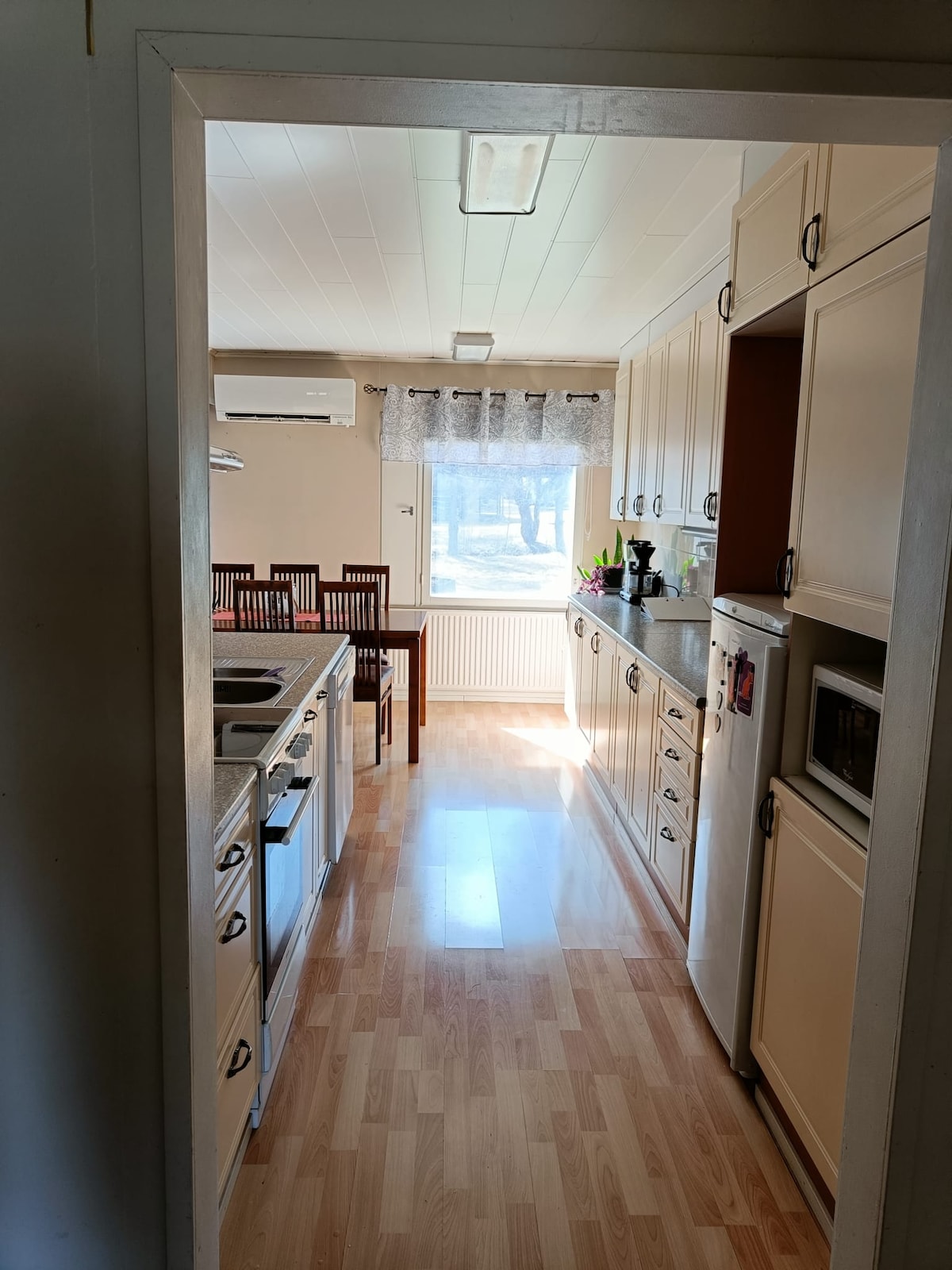 The kitchen features white cabinetry and a spacious layout, with ample counter space. A dining table with wooden chairs is visible, positioned near a sunlit window adorned with sheer curtains. Stainless steel appliances add a modern touch to the welcoming atmosphere.
