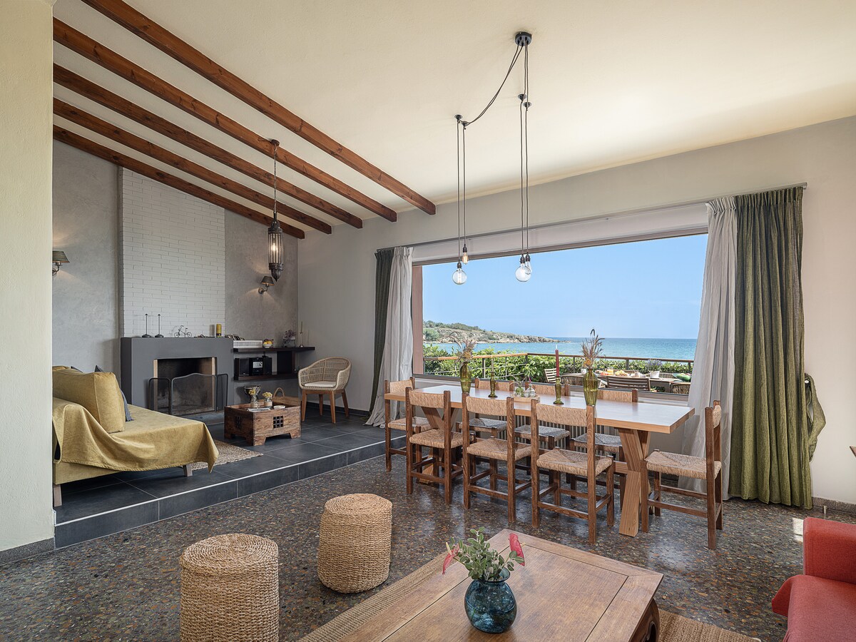 A spacious living area is highlighted by large windows showcasing sea views. A wooden dining table is surrounded by chairs, while a comfortable seating arrangement includes a sofa and decorative poufs. Ceiling beams and pendant lighting enhance the open and airy atmosphere.