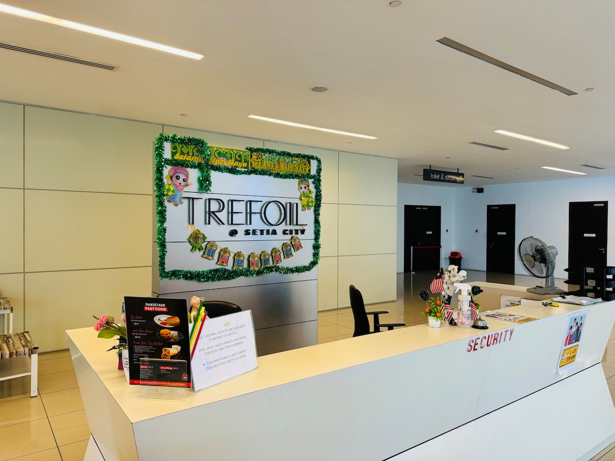 The reception area features a modern design with a sleek front desk. A decorative sign reading 'TREFOIL Setia City' is displayed prominently, surrounded by greenery. The space is illuminated by natural light, and a security desk is positioned nearby, maintaining an organized and welcoming environment.