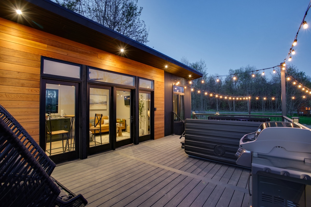 A spacious deck features sliding glass doors leading into the cabin, illuminated by string lights. A hot tub and propane grill are positioned on the wooden surface, complemented by stacking chairs. The natural surroundings provide a serene backdrop for outdoor enjoyment.