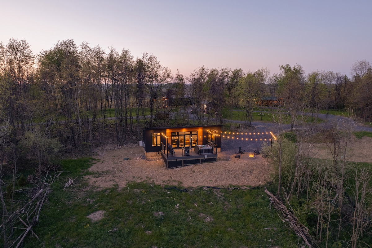 An aerial view showcases the cozy Alpine Abode cabin during twilight, surrounded by trees and natural landscape. Warm string lights illuminate the deck area, while a firepit with chairs is visible nearby, creating a serene setting in the heart of nature.