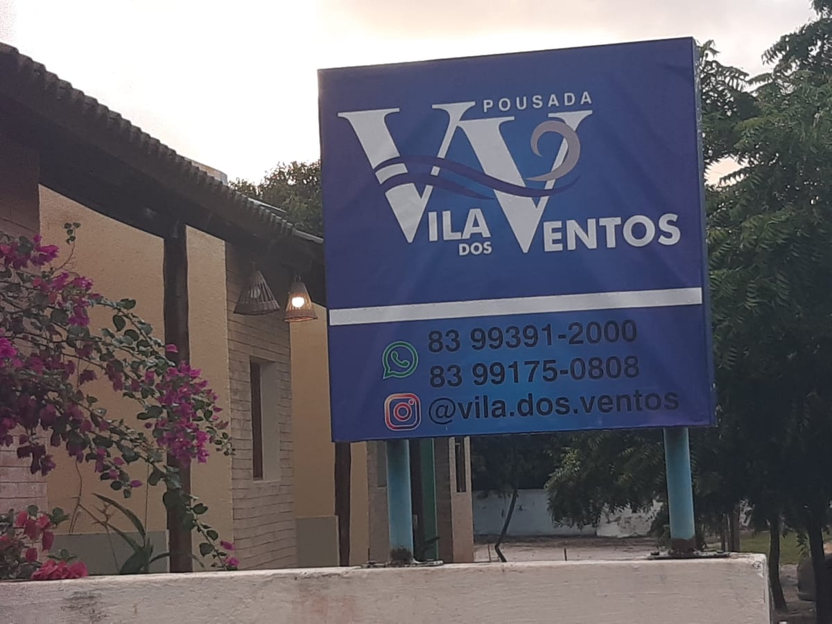 A large sign displaying the name 'Vila dos Ventos' is prominently placed outside the facility. The sign includes contact information and social media handles, surrounded by vibrant green foliage and colorful flowers that enhance the welcoming environment.