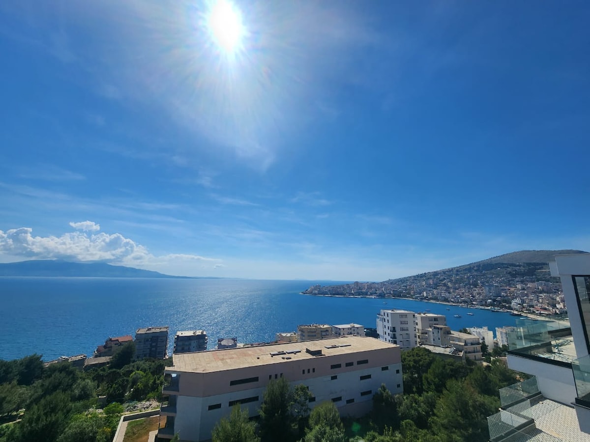 A clear view of the sea and city landscape is captured under a bright blue sky. The sparkling water reflects sunlight, while the coastline features various buildings and greenery, creating a sense of openness and tranquility.