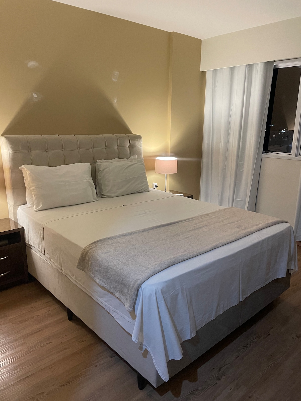 A spacious bed is positioned against a light-colored wall, featuring two soft pillows. A neutral blanket is neatly laid over the bed. A bedside lamp emits a warm glow, while a window with sheer curtains allows for soft light to filter in.