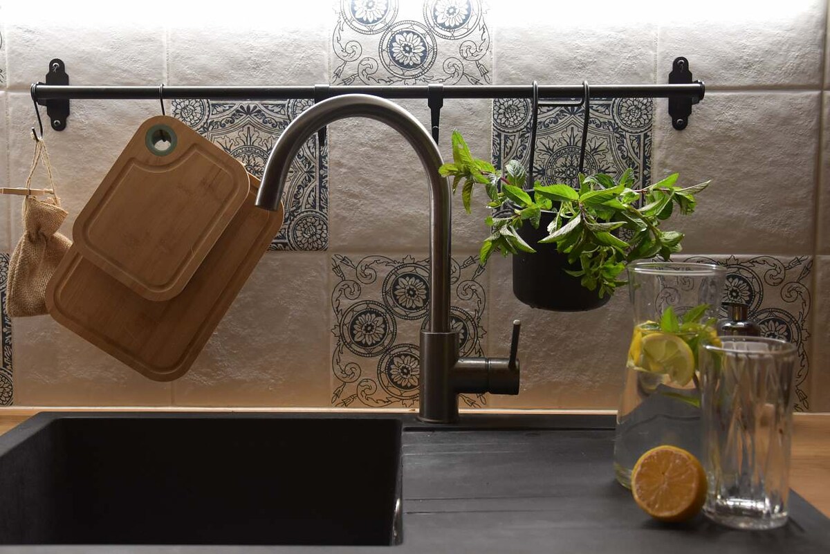 A modern kitchen faucet is mounted above a dark sink, accompanied by two wooden cutting boards hanging from a metal rack. Fresh herbs are displayed in a hanging pot, while a glass pitcher and tall glasses filled with water and lemon sit on the countertop, adding a refreshing touch.