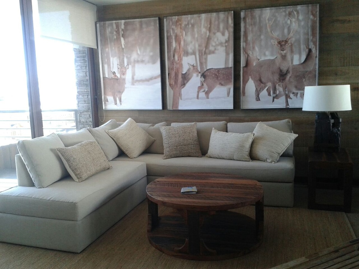 A spacious seating area is highlighted by a large sectional sofa adorned with decorative pillows. A wooden coffee table is placed in front, while three framed images of deer in a snowy landscape are mounted on the wall behind the couch.