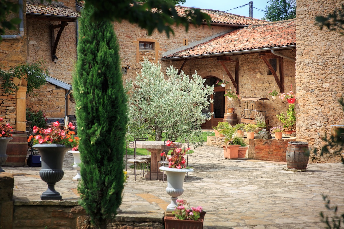 A rustic courtyard is framed by stone walls and a tiled roof. Potted flowers and plants add color around a central table and chairs. Olive trees provide greenery, while terracotta planters and barrels complete the inviting outdoor space.