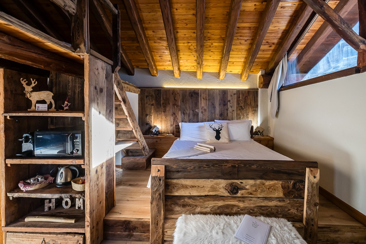 A cozy sleeping area is set within a rustic wooden structure. A spacious bed is positioned under wooden beams, complemented by wall-mounted decor of antlers. Nearby, shelving displays a mini fridge and decorative items, while natural light enters through a window draped with sheer fabric.