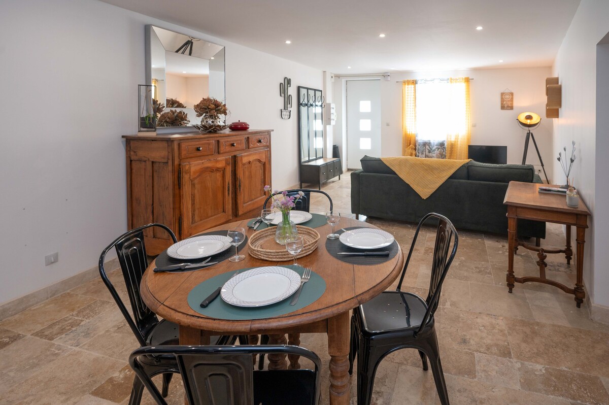 A cozy dining area features a round wooden table set for four, complemented by black metal chairs. A sideboard displays decorative items, with a vase of flowers at the center. Natural light fills the space, highlighting the inviting atmosphere of the open living area.