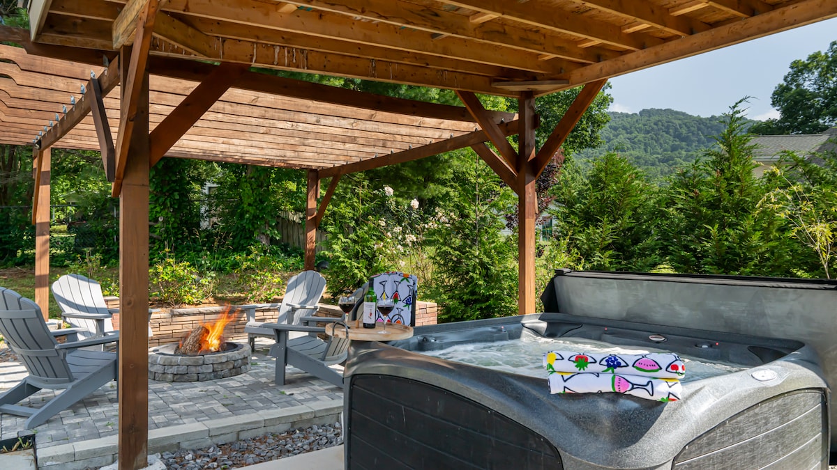 A covered outdoor space features a hot tub next to a stone fire pit, surrounded by lush greenery. Comfortable Adirondack chairs provide seating, and colorful artwork rests on a small table. Warm colors and natural elements create a relaxing gathering area.