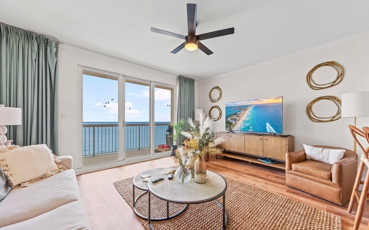 A bright living room features large glass doors opening to ocean views. A comfortable sofa is paired with a brown leather chair. A circular coffee table displays decorative items, while a ceiling fan adds to the airy atmosphere. Warm light filters through the room.