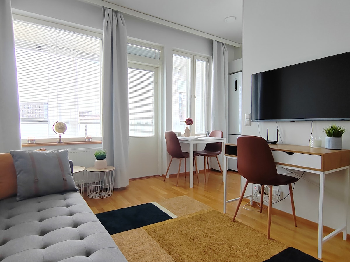 A bright living area is showcased with large windows allowing natural light to fill the space. A plush sofa is complemented by a round side table and a minimalist dining area with a table and two chairs. A mounted television and a small plant add to the decor.