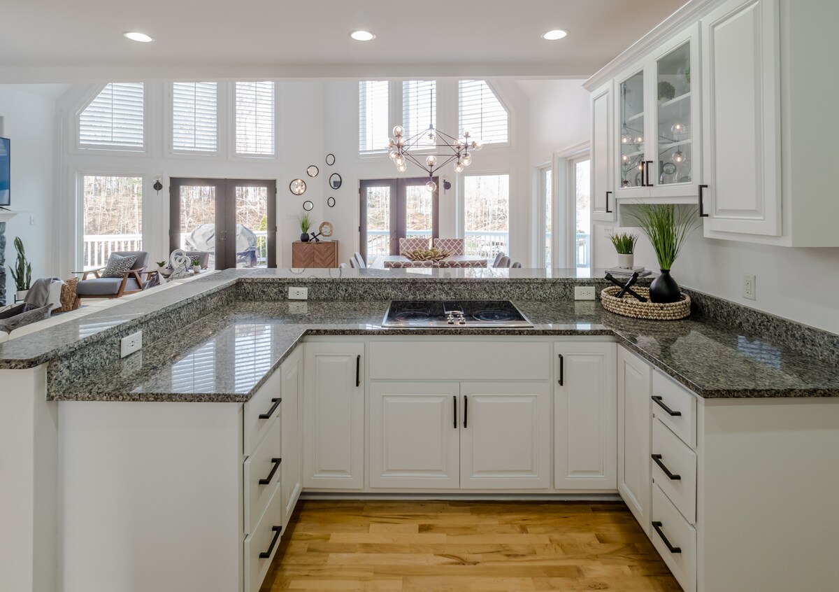 A modern kitchen features a large granite countertop with ample space for meal preparation. White cabinetry offers storage, while natural light floods in through tall windows. A view of the open living area is visible in the background, creating an inviting atmosphere.