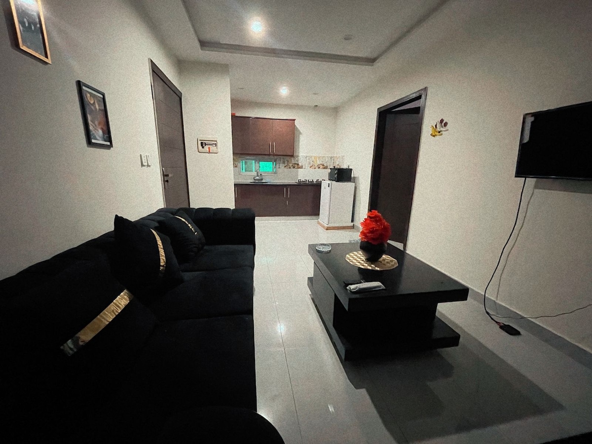 The living area features a plush black sofa paired with a modern coffee table. A small decorative arrangement is placed on the table. The kitchen area is visible in the background, showcasing cabinetry and essential appliances, with bright lighting enhancing the spacious feel.