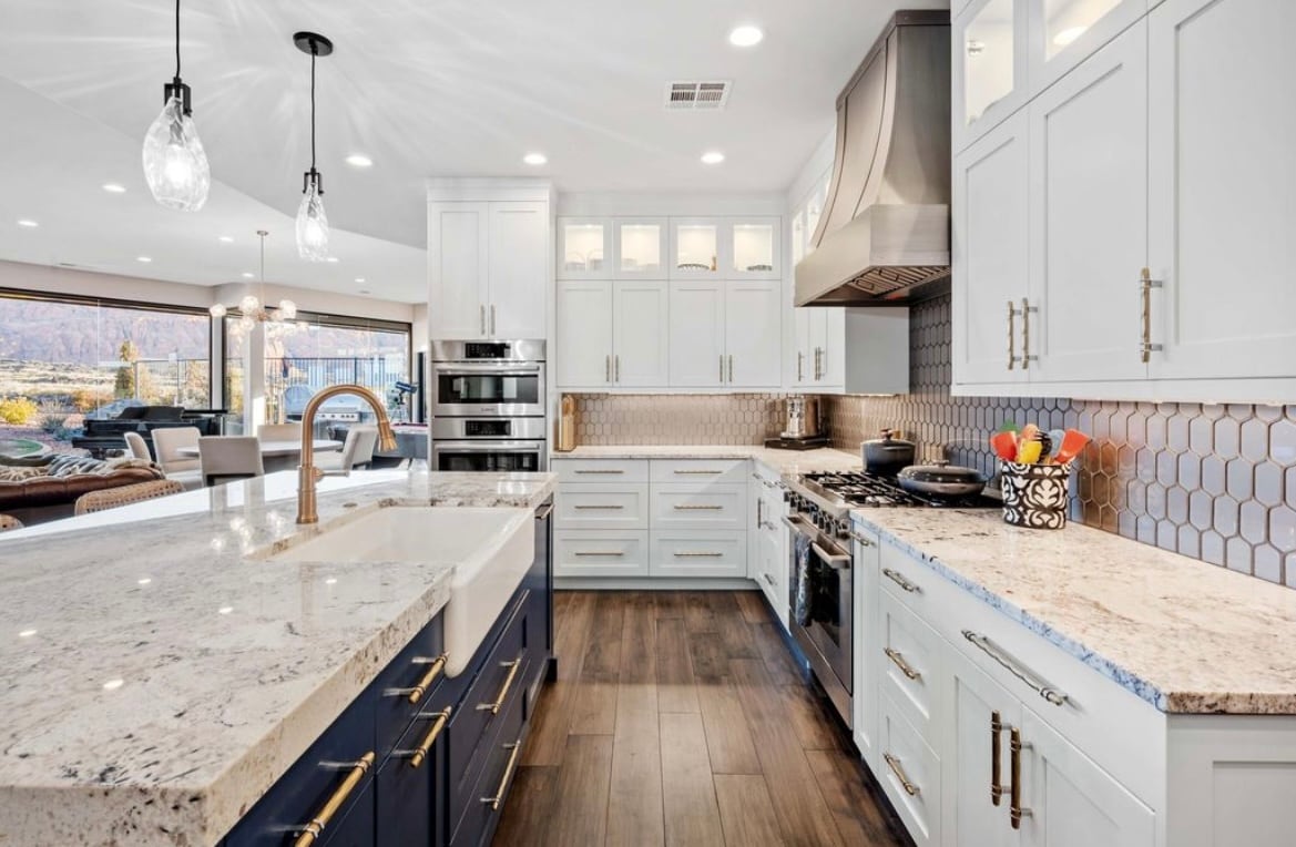 A modern kitchen is presented, featuring white cabinetry and dark blue accents. Natural stone countertops with intricate patterns are visible. A stainless-steel range hood complements the stovetop, while a variety of kitchen appliances are neatly integrated. Large windows offer views of the outdoors, enhancing the spacious feel.