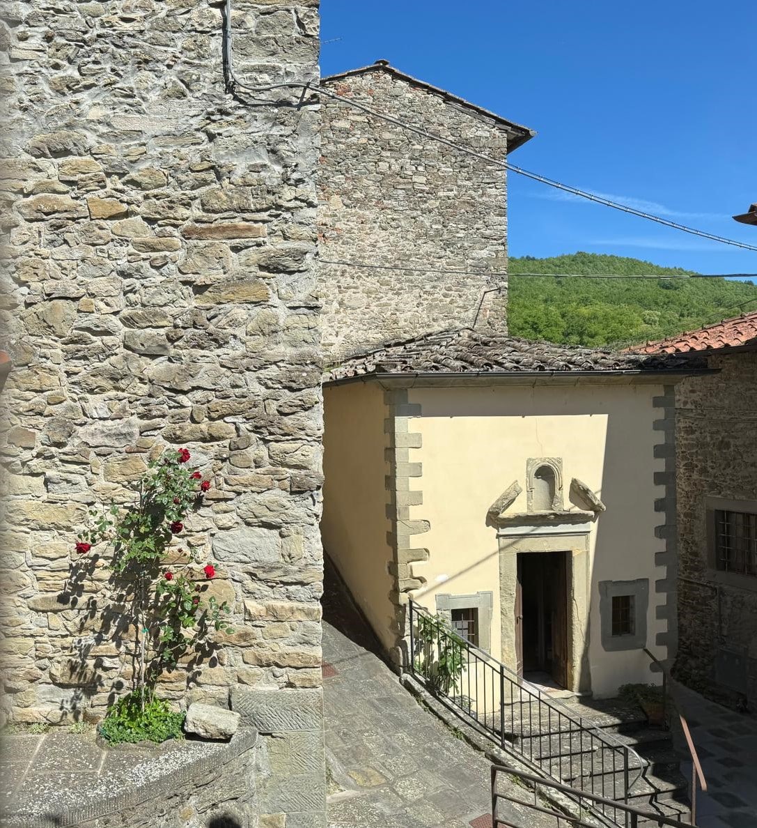 A stone building is visible alongside a winding staircase leading to a quaint entrance. The bright yellow façade of the adjacent structure contrasts with the rugged texture of the nearby stones. A single flowering plant provides a touch of greenery amidst the historical architecture.