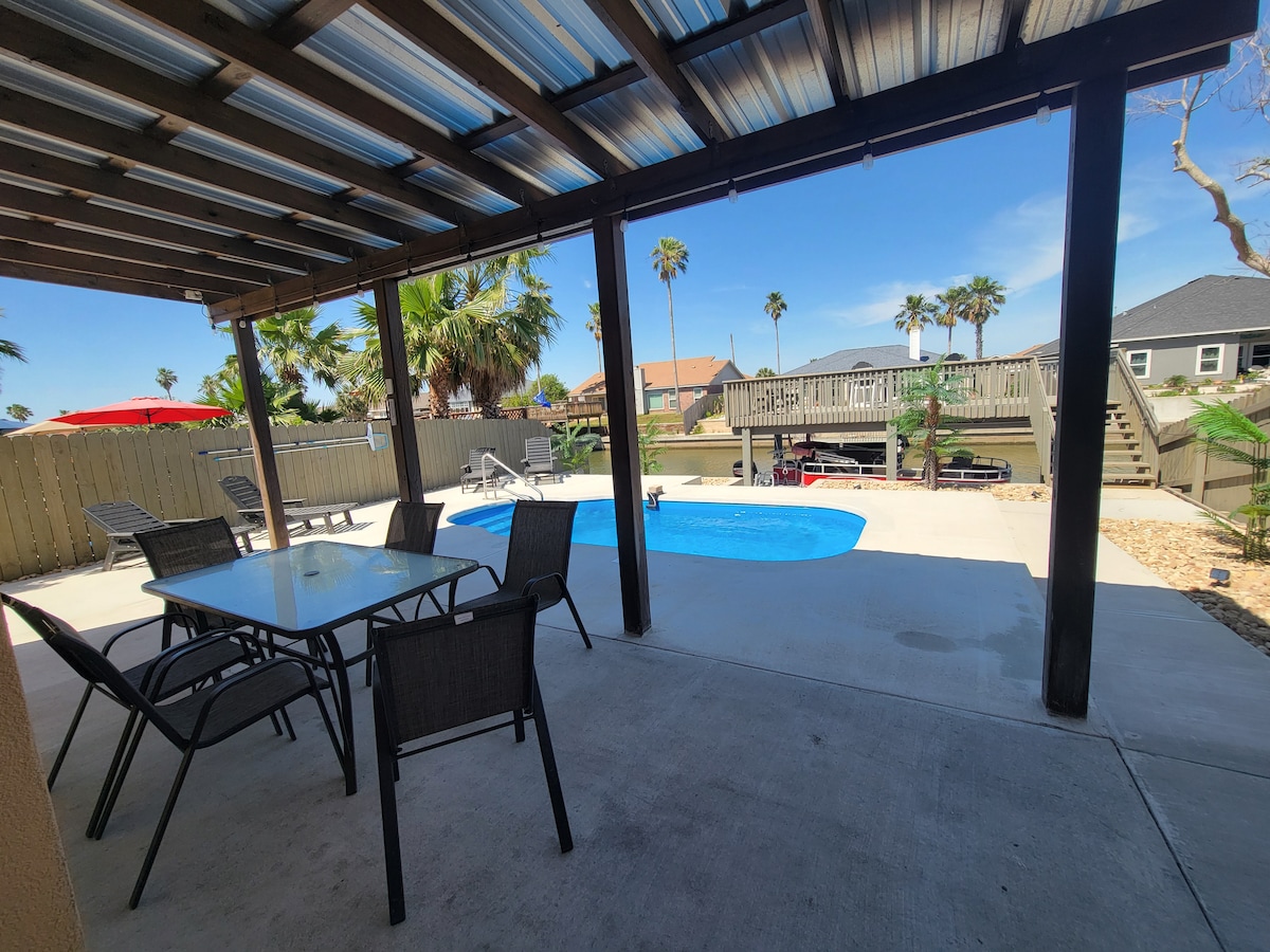 An outdoor dining area is set under a covered patio, featuring a table and six chairs. The private pool is framed by palm trees, with a deck visible in the background. Sunlight brightens the spacious area, highlighting the inviting atmosphere.