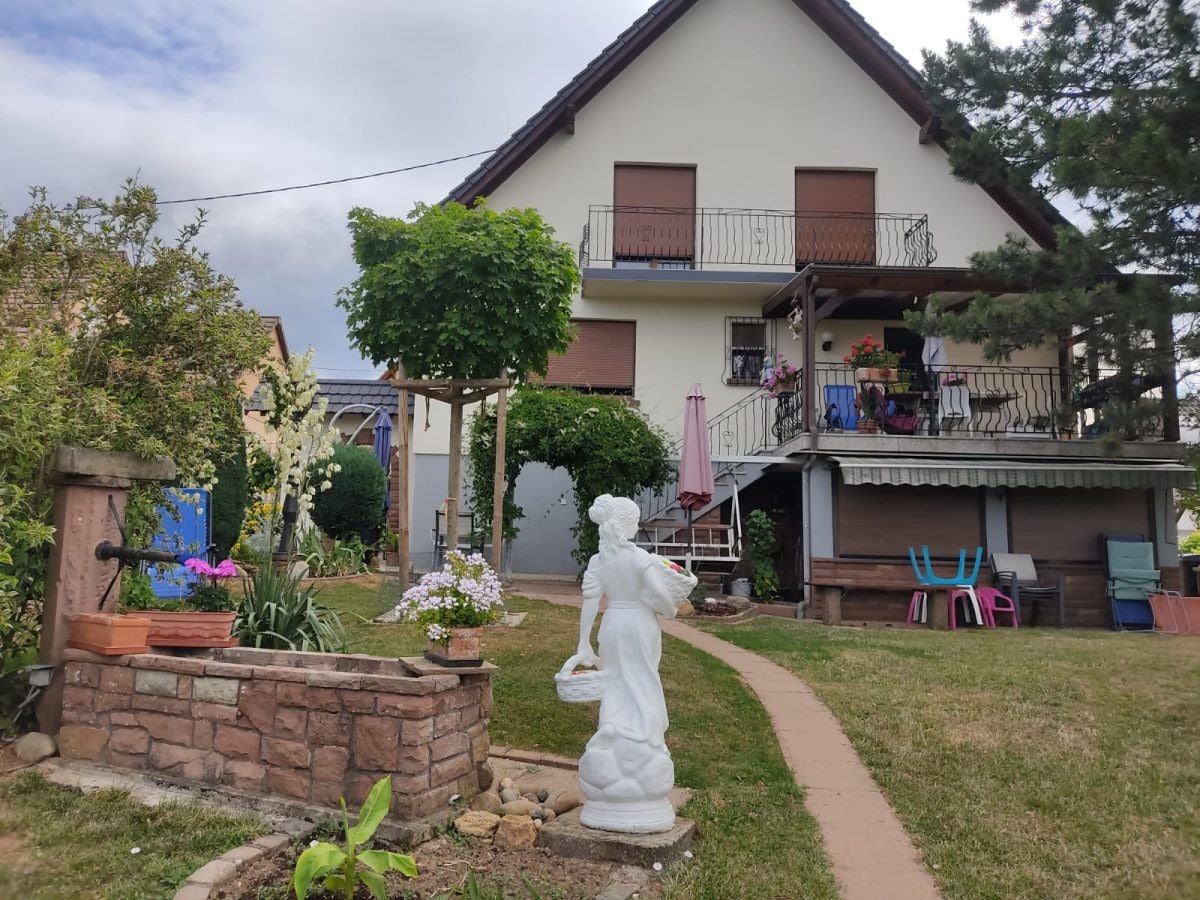 A charming house is positioned amidst a landscaped yard featuring a stone well and a decorative statue. Colorful chairs are arranged on the balcony above, while flower beds bloom alongside a stone pathway that leads to the entrance.