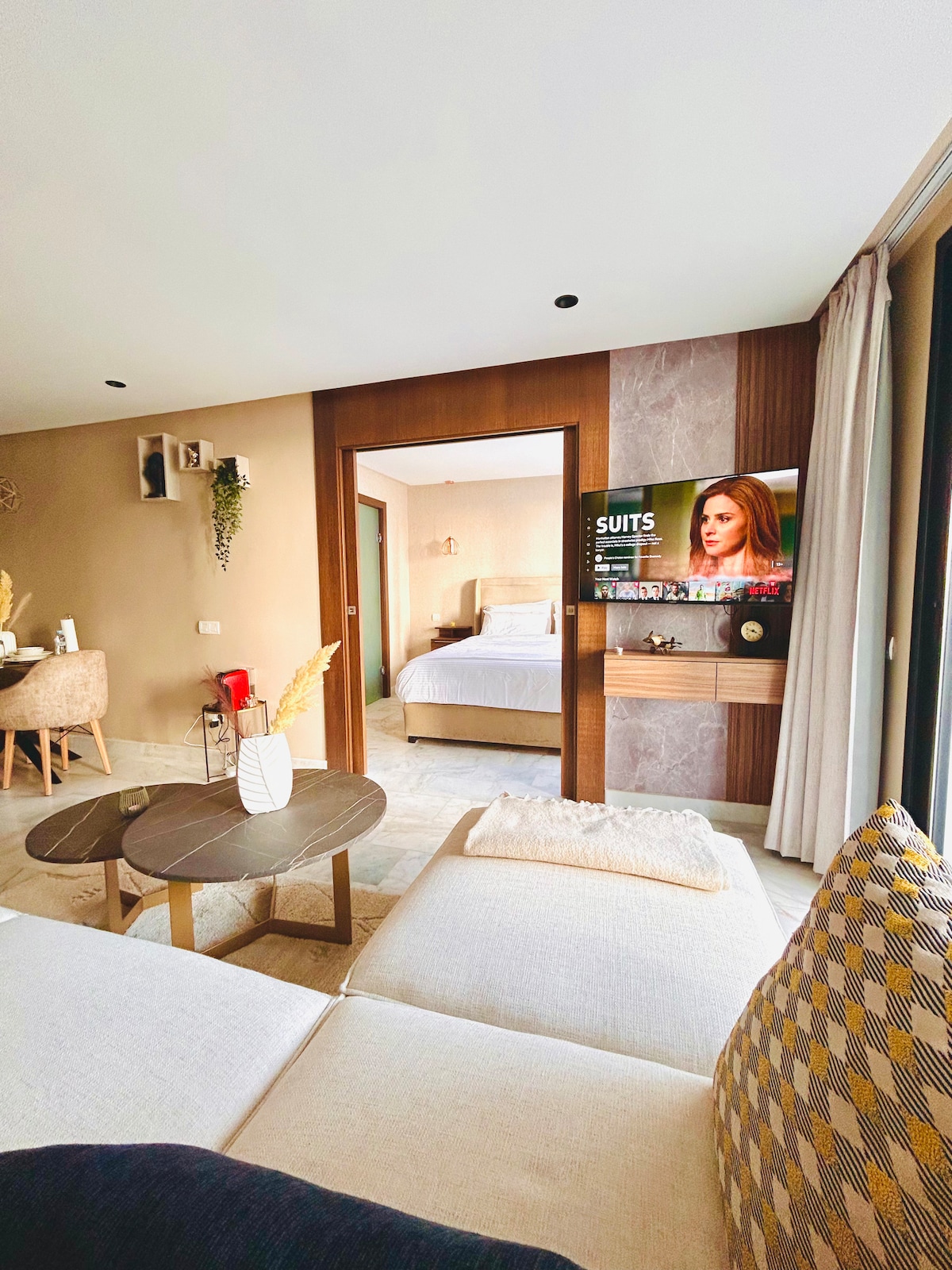A spacious living area features a comfortable sofa with patterned cushions, facing a Smart TV mounted on the wall. The room is bathed in natural light from large windows, while a dining area is visible in the background, leading to a separate bedroom.