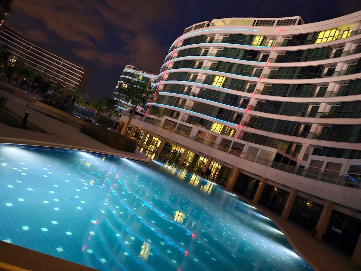 An elegantly designed residence complex is highlighted, showcasing multiple curved buildings illuminated at night. The inviting outdoor pool reflects the building's lights, with a clear blue surface adorned with subtle tile patterns. Surrounding palm trees add a touch of greenery to the scene.