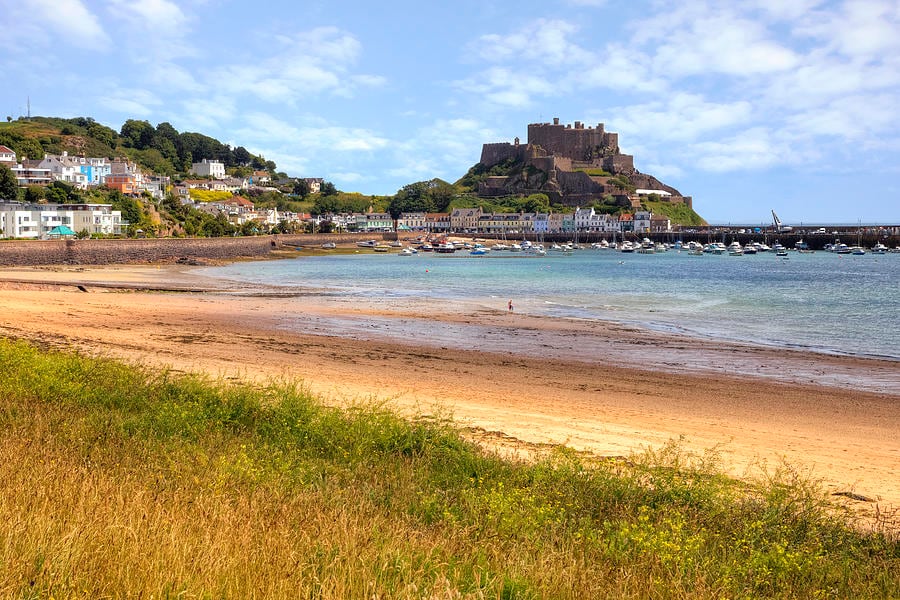 The image captures a sandy beach stretching along the coastline, with gentle waves lapping at the shore. In the background, a castle is perched on a hill, surrounded by greenery and buildings, while boats are anchored in the harbor. A clear blue sky adds to the scene.